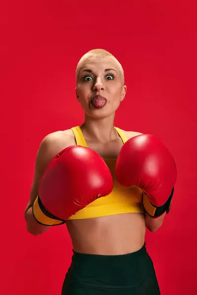 Portrait of attractive young woman in boxing gloves grimacing looking at camera against red studio background. Concept of sport, hobby, active-lifestyle, self expression, human emotions.