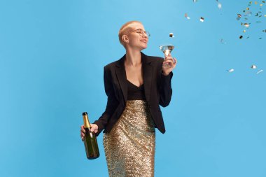 Zarif giyimli genç bayan elinde şampanya şişesi ve alkol tutarken konfetiler mavi stüdyonun arka planında uçuyor. Cuma havası, kutlama, tatiller ve partiler, duygular.