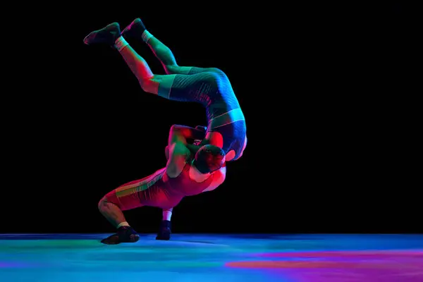 Two young man, professional sportsman of freestyle wrestling in uniform fighting against black background in mixed neon lights. Concept of motion, action, combat sports, strength and power, movement.