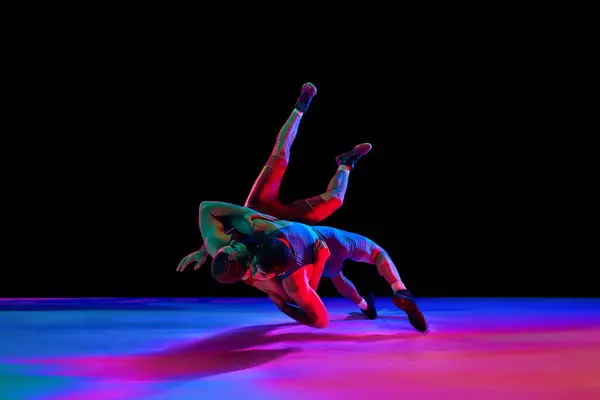 Greco-Roman, freestyle wrestling. Two sportsman, fighting in red and blue uniform in action against black background in mixed neon lights. Concept of motion, action, combat sports, strength and power.