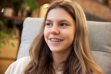 Diş telleriyle gülümseyen genç ve çekici bir kızın portresi. Koltukta oturuyor ve başka tarafa bakıyor. Gençlik kavramı, sağlık sigortası, mükemmel gülümseme, diş sorunları.