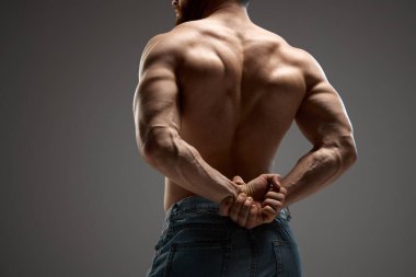 Kaslı bir erkek, model elleri arkadan kenetlenmiş. Mab kot pantolon giyiyor, gri arka planda poz veriyor. Güzellik ve moda kavramı, erkeklik, vücut geliştirme, tarz.