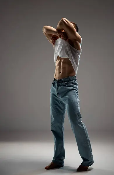 Full-length photo of man, model with an athletic and muscular body taking off his T-shirt in motion against grey studio background. Concept of beauty and fashion, masculinity, bodybuilding, style.