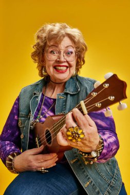 Ukulele çalan, sarı arka planda kameraya bakan heyecanlı, modaya uygun yaşlı kadın portresi. Daha yaşlı yetişkinlerin, modern hayatın aktif yaşlılarının, moda ve tarzın güçlendirilmesi kavramı.