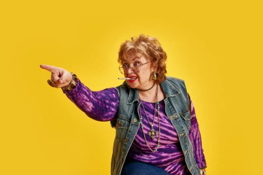 Ağzında sigara olan yaşlı bir kadın bir yeri göstererek kot ceket ve kravat boyası tişört giyiyor. Direktif duygular. Daha yaşlı yetişkinlerin, modern hayatın aktif yaşlılarının yetkilendirilmesi kavramı.