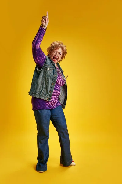 Vibrant, energetic elderly woman in modern clothes with jewelry looks at camera and poses against yellow background. Concept of empowerment of older adults, active seniors in modern life, fashion.