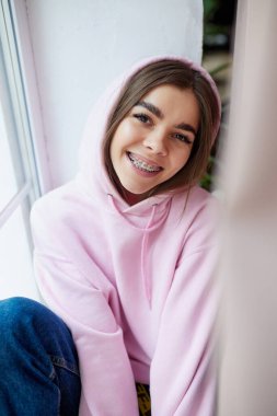 Pencere kenarında pembe kapüşonlu ve kameraya bakıp gülümseyen genç, pozitif kadın portresi. Yukarıdan bak. Güzellik ve ilaç kavramı, boş zaman, moda.