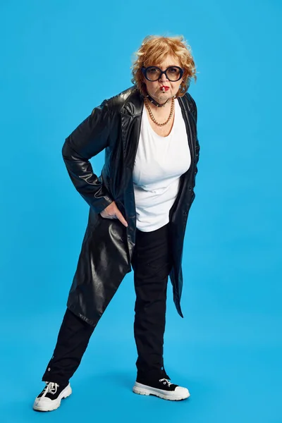 Full length portrait of cool attitude mature woman, pensioner with cigarette posing against blue studio background. Concept of fashionable older adults, empowerment of older adults. Ad