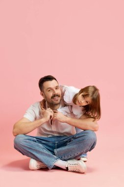 Genç bir adam oturuyor ve küçük bir kız onunla oynuyor. Pembe arka planda baba ve kız çocukları arasında sıcak ve sevimli bir ilişki var. Uluslararası Mutluluk Günü kavramı, çocukluk ve ebeveynlik.