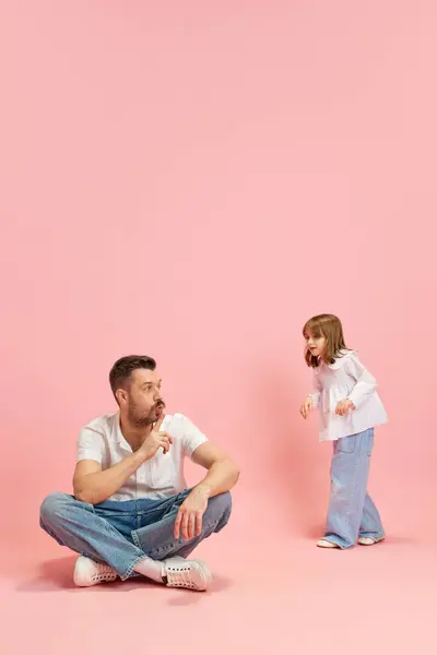Seated man, dad gesturing to keep silence to walking so loud and speaking child against pink pastel background. Concept of International Day of Happiness, childhood and parenthood, emotions. Ad