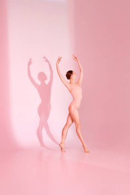 Pastel Bale. Kadın zarif bir şekilde pembe mayoyla pembe arka plana poz veriyor. Çıplak ayaklı silueti, zarif hareketlerini yansıtan gölgeler bırakıyor. Bale, zarafet, güzellik kavramı.