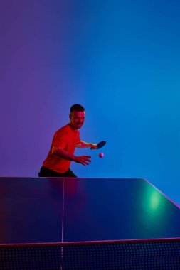 Spor üniformalı masa tenisçisi topa yoğun bir şekilde vuruyor. Kırmızı neon ışıklı, filtreli mor-mavi arka plan. Dinamik jel portresi. Spor anlayışı, sağlıklı yaşam tarzı. Ad