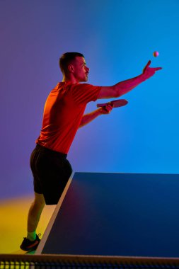 Yan görüş. Masa tenisinde top oynayan genç atlet, arka plandaki neon ışıkta yüzen topa bakıyor. Dinamik jel portresi. Profesyonel spor anlayışı, enerji. Ad