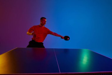 Hızlı masa tenisi oynayan atletik bir adamın fotoğrafı neon ışıkta mavi-mor arka plana karşı. Uzayı kopyala Dinamik jel portresi. Profesyonel spor anlayışı, sağlıklı yaşam tarzı. Ad