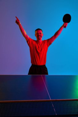 Muzaffer genç adam, masa tenisçisi kollarını neon ışığında kaldırarak mor-mavi arka plana karşı kutlama yapıyor. Dinamik jel portresi. Profesyonel spor kavramı, kazan. Ad