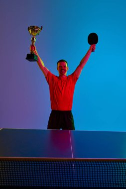 Kırmızı tişörtlü neşeli masa tenisi oyuncusu el kaldırdı ve arka planda neon ışıkta bardağı tuttu. Dinamik jel portresi. Profesyonel spor anlayışı, sağlıklı yaşam tarzı, kazanan.