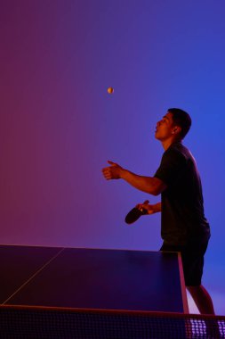 Genç adam, oyuncu neon ışıklı masa tenisi maçında pembemsi arka planda topa vurmaya hazır. Dinamik jel portresi. Profesyonel spor anlayışı, sağlıklı yaşam tarzı ve eğlence. Ad