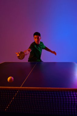 Yeşil spor giyimli, odaklanmış masa tenisçisi, topa göz kulak oluyor, eğimli arka plana karşı ön atış için hazırlanıyor. Neon. Dinamik jel portresi. Profesyonel spor anlayışı, enerji. Ad
