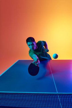 Masa tenisinde, neon ışıkta, sıcak sarı arka planda, Asyalı bir adam konsantrasyonunu koruyor. Dinamik jel portresi. Profesyonel spor anlayışı, sağlıklı yaşam tarzı ve eğlence. Ad