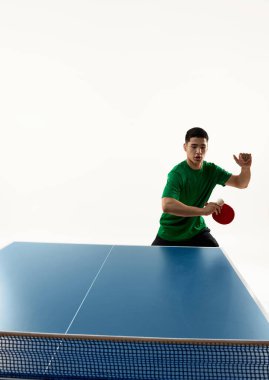 Formda Asyalı bir adam, yeşil spor üniformalı masa tenisi oyuncusu beyaz arka planda mükemmel vuruş için topa odaklanmış. Hareket var. Spor, hobi, yaşam tarzı, maç, zafer, şampiyonluk kavramı. Ad