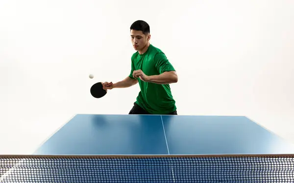 Focused table tennis serve from Asian player in vibrant green sportswear in motion against white studio background. Frozen moment. Concept of sport, hobby, lifestyle, match, victory, championship.