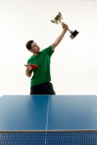 Player in victory pose holds cup and racquet at table tennis match against white studio background. Power and strength. Concept of sport, hobby, lifestyle, match, victory, championship. Ad