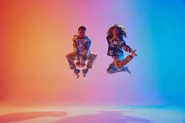 Energetic positive pair, dancers leaping in neon colorful illumination suspended in mid-air against gradient background. Concept of movement, energy, dance battles. Dynamic gel portrait