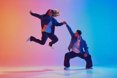 Kadın dansçının havadaki dinamik çekimi, erkek dansçının çömelmesi ve neon ışıkta renkli arkaplanda el ele tutuşması. Gençlik kültürü, müzik, yaşam tarzı, tarz, moda kavramı.