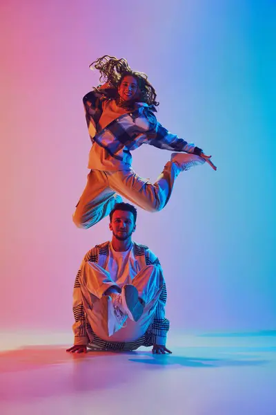 Dance duet. Woman jumps over man crouching in radiant neon glow. Dynamic dance move and joyful energy. Concept of movement, music, freestyle, dance battles, motion. Dynamic gel portrait