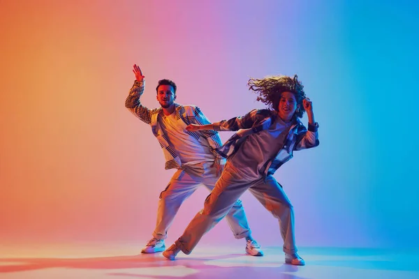 Dynamic shot of athletic man and woman, hip-hop dancers learning new dance routine against gradient studio background in neon light. Concept of movement, energy, dance battles. Dynamic gel portrait