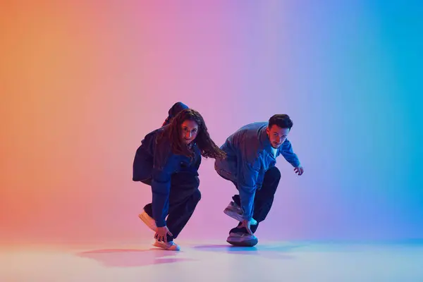 Two dancers performing sync hip-hop moves dressed in denim clothes against gradient background in neon light. Concept of youth culture, music, lifestyle, style and fashion, action. Gel portrait.