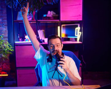 Mutlu ve heyecanlı yayıncının portresi, blog yazarı renkli, neon ışıklı çalışma alanında ses kaydediyor, ev stüdyosunda. Sosyal medya ve akıllı çalışma, yaşam tarzı ve çevrimiçi gençlik kavramı.