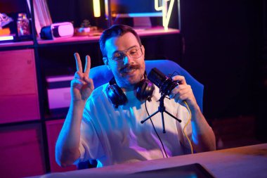 Genç bir adam, blogcu jesti, podcast 'i kaydederken gösteri işareti, ev stüdyosundaki ruh haliyle aydınlatılmış çalışma alanında. Sosyal medya ve akıllı çalışma, yaşam tarzı, yayın platformu olan gençlik kavramı.