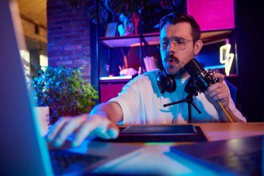 Neon ışıklı çalışma alanındaki stüdyo mikrofonuna konuşurken çoklu görev podcaster ayarlama ekipmanı. Sosyal medya ve akıllı çalışma, yaşam tarzı, yayın platformu olan gençlik kavramı.