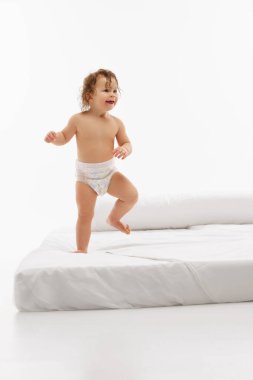 Neşeli küçük bir çocuk, küçük bir çocuk, ıslak saçlı ayakta duran bir kız parlak beyaz stüdyo arka planına karşı yatakta oynuyor. Çocukluk kavramı, annelik, yaşam, doğum. İlan için alanı kopyala
