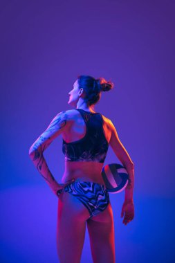 Arkadan bakan genç atlet kadın portresi, plaj voleybolcusu topla mavi-mor arka planda neon ışıkta poz veriyor. Spor anlayışı, hareket, aktif ve sağlıklı yaşam tarzı, güç.