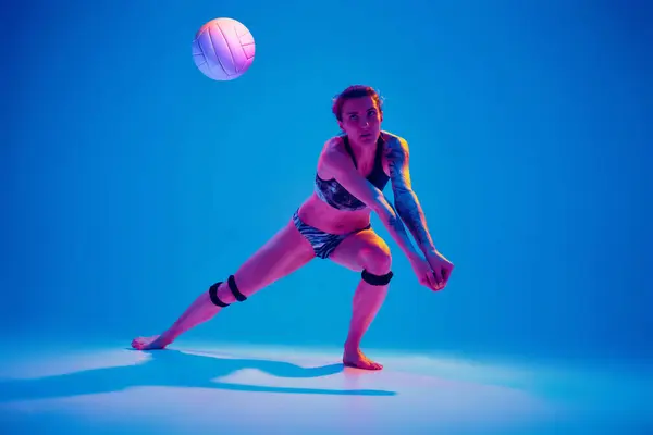 Sportswoman crouched in ready position to hitting ball, make low pass against gradient blue background in pink neon light. Concept of sport, movement, active and healthy lifestyle, power and strength.