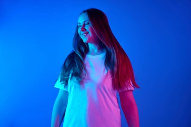 Beyaz tişörtlü mutlu, keyifli bir kadının portresi. Pembe neon ışıklı, gradyan mavi stüdyo arka planına karşı poz veriyor. Güzellik ve moda kavramı, kendini ifade etme, insani duygular. Ad