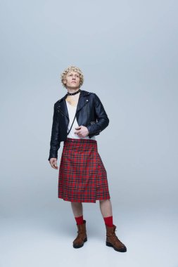 İskoç eteği ve deri ceketli, gri stüdyo arka planında poz veren yakışıklı, kıvırcık saçlı genç adamın tam boy portresi. Moda birleşimi, sanat fotoğrafçılığı, stil, kendini ifade etme kavramı.