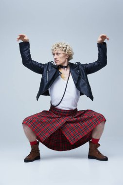 İskoç eteği ve deri ceketli adam kollarını kaldırarak poz veriyor. Gri stüdyo arka planına geleneksel ve punk tarzının bir karışımını somutlaştırıyor. Moda birleşimi, sanat, eşsizlik, kendini ifade etme kavramı. Ad