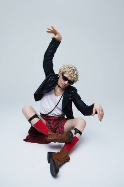 Deri ceketli ve ekose etekli bir adam dinamik dans pozunda oturan güneş gözlüklü ve botlu gri stüdyo arka planına karşı. Moda birleşimi, sanat fotoğrafçılığı, stil, nonoş, kendini ifade etme kavramı.