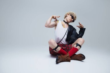 Lotus pozunda oturan şık genç adam İskoç eteği ve gri arka planda modern deri ceket giyiyor. Çağdaş ve gelenekler. Moda birleşimi, sanat, eşsizlik, kendini ifade etme kavramı. Ad