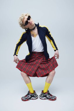 Ekose etekli neşeli bir adam ve spor ayakkabılı ve sarı çoraplı eski eşofman üstü dans eden gri stüdyo arka planında. Moda birleşimi, sanat fotoğrafçılığı, stil, eşsizlik, kendini ifade etme kavramı.