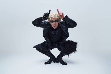 Siyah giysili ve çizmeli genç adam, gri stüdyo geçmişine karşı hareketli ve isyankar bir tavırla taş atıyor. Moda birleşimi, sanat fotoğrafçılığı, stil, nonoşluk, eşsizlik kavramı. Ad