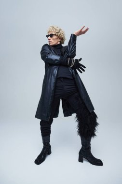 Gri stüdyo arka planına poz veren uzun siyah ceketli, eldivenli ve topuklu çizmeli bir gencin tam boy portresi. Moda birleşimi, sanat fotoğrafçılığı, eşsizlik, kendini ifade etme kavramı. Ad