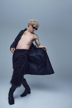 Gri stüdyo arka planına karşı çıplak ve modaya uygun giysiler içinde poz veren etkileyici genç bir adam. Moda birleşimi, sanat fotoğrafçılığı, tarz, nonoşluk, eşsizlik, kendini ifade etme kavramı. Ad