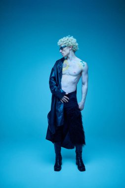 Neon ışıklı stüdyoda mavi arka planda poz veren atletik, genç bir adamın tam boy portresi. Moda birleşimi, sanat fotoğrafçılığı, tarz, nonoşluk, eşsizlik, kendini ifade etme kavramı. Ad