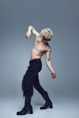 Yarı çıplak genç adam modern pantolonlar ve yüksek topuklu ayakkabılar giyerek gri stüdyo arka planında dans ediyor. Moda birleşimi, sanat fotoğrafçılığı, tarz, nonoşluk, eşsizlik, kendini ifade etme kavramı. Ad