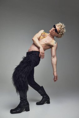 Gri stüdyo arka planında siyah pantolon ve yüksek topuklu ayakkabı giyen çıplak vücutlu genç yakışıklı bir adam. Moda birleşimi, sanat fotoğrafçılığı, tarz, nonoşluk, eşsizlik, kendini ifade etme kavramı. Ad