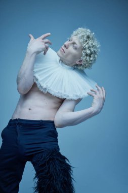 Kıvırcık saçlı, avangart görünümlü bir adam klasik bir dokunuşla ve neon ışıkta mavi arka plana karşı anlamlı ellerle. Moda birleşimi, tarz, nonoş, eşsizlik, kendini ifade etme kavramı.